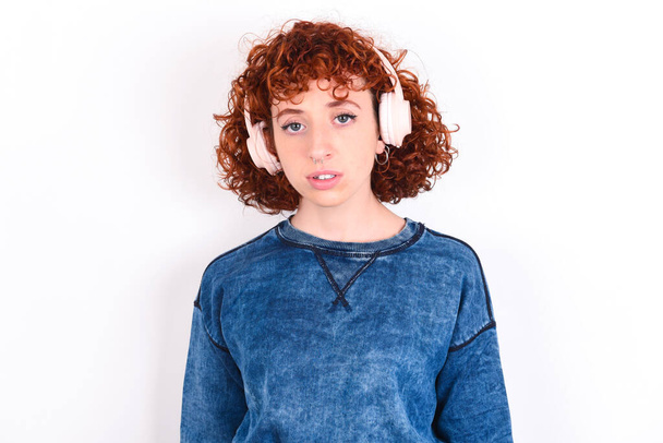Poważny niezadowolony młoda kaukaska kobieta rude włosy noszenie niebieski T-shirt na białym tle wygląda zaskoczony na aparat jest zły nosi słuchawki stereo słucha muzyki podczas spaceru na ulicy - Zdjęcie, obraz