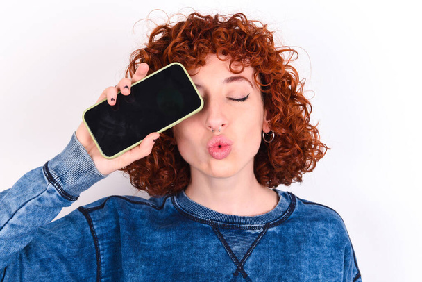 Adorável jovem caucasiano mulher ruiva vestindo azul T-shirt sobre fundo branco segurando dispositivo moderno cobrindo o olho com lábios amuados - Foto, Imagem