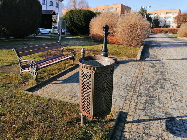 Sentado na praça. Um banco no meio do parque na praça, uma lata de lixo de metal e um hidrante em uma área pavimentada entre a geenery.  - Foto, Imagem