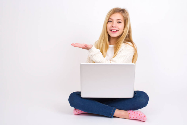Ein kaukasisches Teenie-Mädchen, das mit Laptop in Lotusposition auf weißem Hintergrund sitzt, sagt: Wow, wie aufregend es ist, hat erstaunte Mienen, zeigt etwas auf einer leeren Fläche mit Handfläche. Werbekonzept. - Foto, Bild