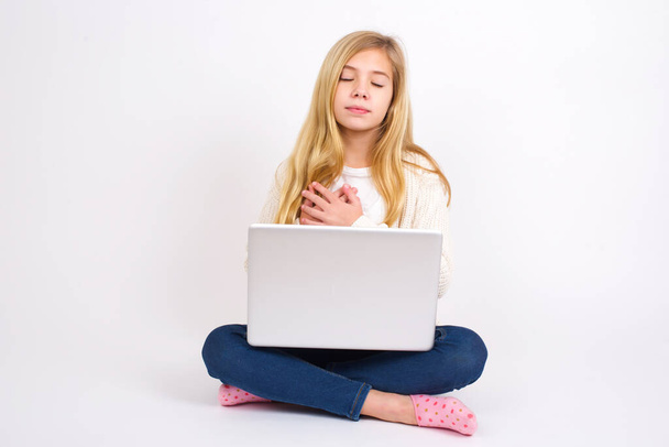 caucasian teen girl sitting with laptop in lotus position on white background  closes eyes and keeps hands on chest near heart, expresses sincere emotions, being kind hearted and honest. Body language and real feelings concept. - Photo, Image