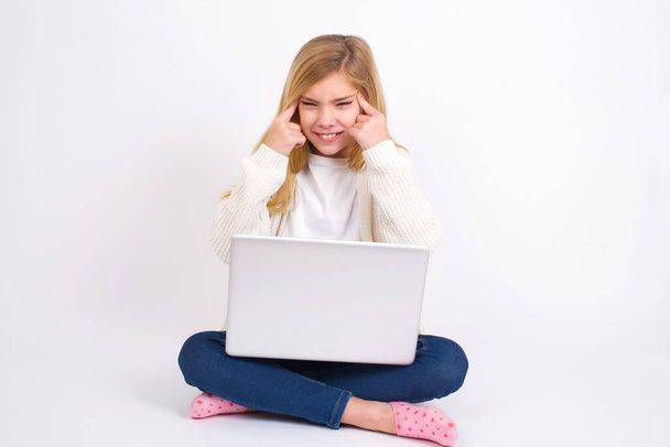 Καυκάσιος έφηβος κορίτσι κάθεται με φορητό υπολογιστή σε θέση λωτού σε λευκό φόντο επικεντρώνεται σκληρά σε μια ιδέα με μια σοβαρή ματιά, σκέψης και με τα δύο δάχτυλα δείκτη δείχνει προς το μέτωπο. - Φωτογραφία, εικόνα