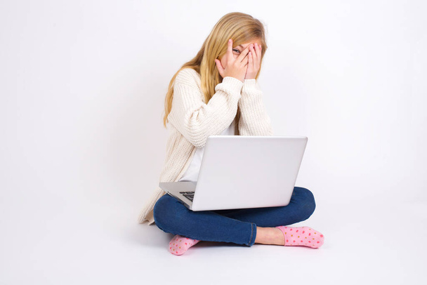 Kaukaska nastolatka siedzi z laptopem w pozycji lotosu na białym tle pokrywając twarz rękami i wypatrując jednym okiem między palcami. Boję się czegoś lub kogoś.. - Zdjęcie, obraz