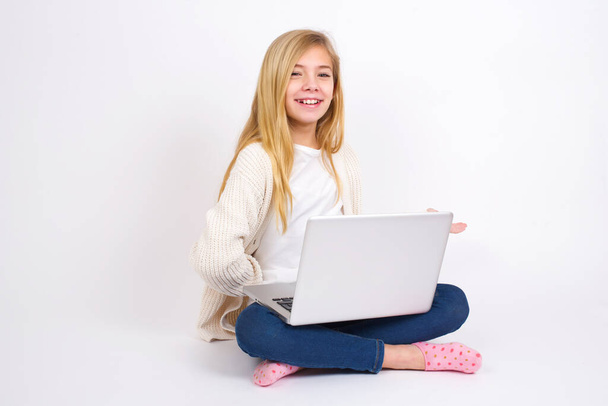 Porträt eines kaukasischen Teenie-Mädchens, das mit Laptop in Lotusposition auf weißem Hintergrund sitzt und den Arm zur Begrüßung ausstreckt. - Foto, Bild