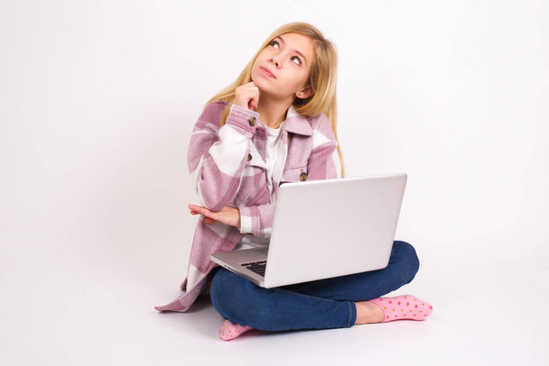 Expressões faciais e emoções. Lindo caucasiano menina adolescente sentada com laptop em posição de lótus no fundo branco segurando a mão sob sua cabeça, tendo olhar duvidoso. - Foto, Imagem