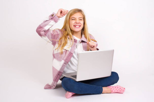 Ελκυστική Όμορφη καυκάσιος έφηβος κορίτσι κάθεται με φορητό υπολογιστή στη θέση του λωτού σε λευκό φόντο γιορτάζει μια νίκη punching στον αέρα με γροθιές του και ένα ακτινοβόλο χαμόγελο toothy. - Φωτογραφία, εικόνα