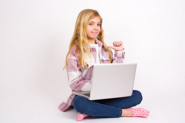Крупним планом весела красива біла дівчина-підліток сидить з ноутбуком в положенні лотоса на білому тлі виглядає радісно, задовільно і впевнено, вказує на себе з великим пальцем
. - Фото, зображення