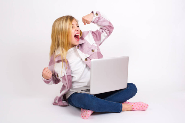 Çekici Kafkas genç kız dizüstü bilgisayarı ile beyaz arka planda oturmuş zaferi kutluyor yumruklarıyla havayı yumrukluyor ve dişlek gibi gülümsüyor. - Fotoğraf, Görsel