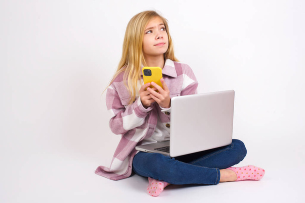 Retrato de bela menina adolescente caucasiana sentado com laptop em posição de lótus no fundo branco com olhar sonhador, pensando ao segurar smartphone. Tenta escrever uma mensagem. - Foto, Imagem