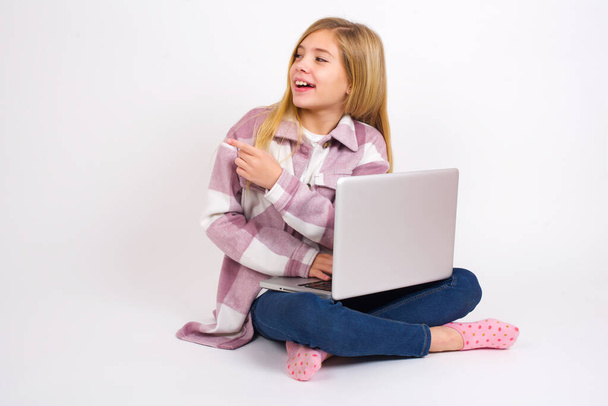 Feliz sorridente alegre Bela menina adolescente caucasiana sentada com laptop em posição de lótus no fundo branco olhando e apontando para o lado com a mão. Espaço de cópia e conceito de publicidade. - Foto, Imagem