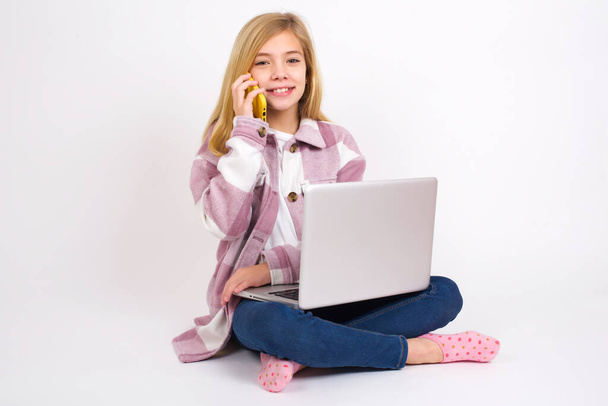 Retrato de una muchacha adolescente caucásica hermosa sonriente sentada con el ordenador portátil en posición del loto en fondo blanco que habla en el teléfono móvil. Concepto de negocio, confianza y comunicación. - Foto, Imagen