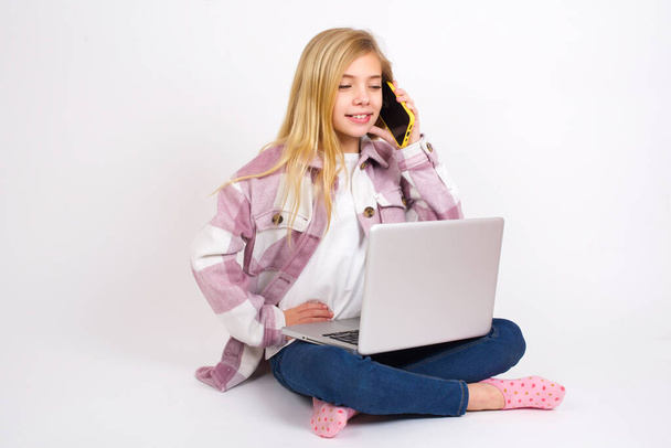 Портрет успішної радісної красивої дівчини-підлітка, яка сидить з ноутбуком в позі лотоса на білому тлі, розмовляючи на мобільному телефоні з другом. Концепція способу життя та спілкування
 - Фото, зображення