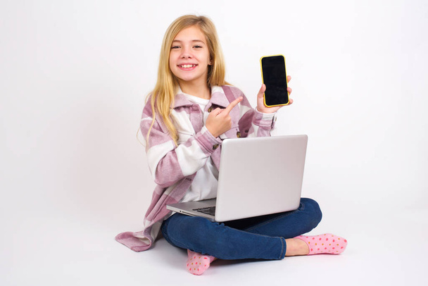 Χαμογελώντας Όμορφη καυκάσιος έφηβος κορίτσι κάθεται με φορητό υπολογιστή σε θέση λωτού σε λευκό φόντο δείχνει και δείχνει σε άδεια οθόνη του τηλεφώνου. Έννοια διαφήμισης και επικοινωνίας. - Φωτογραφία, εικόνα