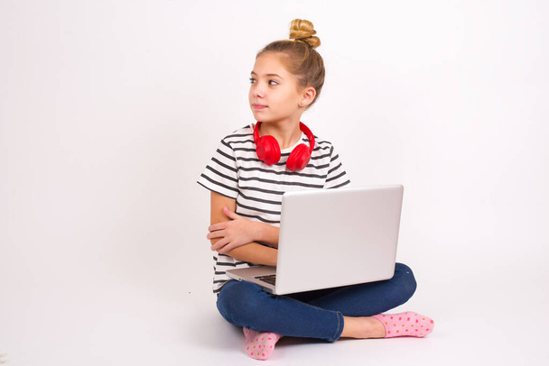 Красивая кавказская девушка-подросток сидит с ноутбуком в позе лотоса на белом фоне держит руки скрещенными над грудью выглядит счастливо в сторону - Фото, изображение
