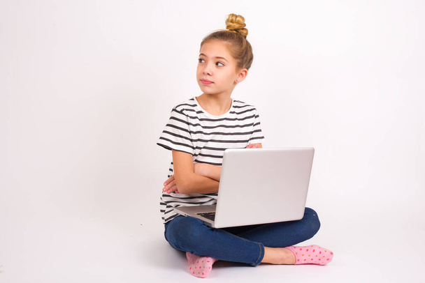 Очаровательная задумчивая красивая кавказская девушка-подросток, сидящая с ноутбуком в позе лотоса на белом фоне, стоит со сложенными руками, сконцентрированная где-то с задумчивым выражением думает, что делать - Фото, изображение