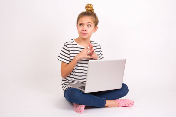 Piękny biały nastolatek dziewczyna siedzi z laptopem w pozycji lotosu na białym tle wieżyczki palce i wygląda tajemniczo na bok ma wielki zły plan w umyśle - Zdjęcie, obraz