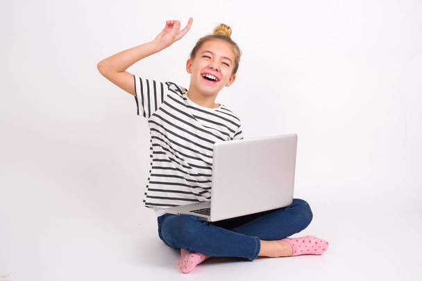 Photo of upbeat Beautiful caucasian teen girl sitting with laptop in lotus position on white background has fun and dances carefree wear being in perfect mood makes movements. Spends free time on disco party - Fotó, kép