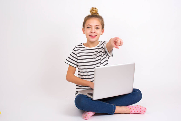 Ενθουσιασμένη θετική Όμορφη καυκάσιος έφηβος κορίτσι κάθεται με φορητό υπολογιστή σε θέση λωτού σε λευκό φόντο σημεία δείκτη απευθείας σε σας, βλέπει κάτι πολύ αστείο. Καταπληκτικό. - Φωτογραφία, εικόνα