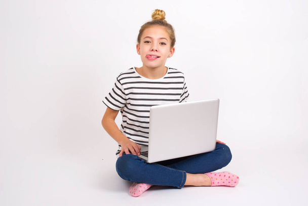 Funny Beautiful caucasian teen girl sitting with laptop in lotus position on white background makes grimace and crosses eyes plays fool has fun alone sticks out tongue. - Photo, Image