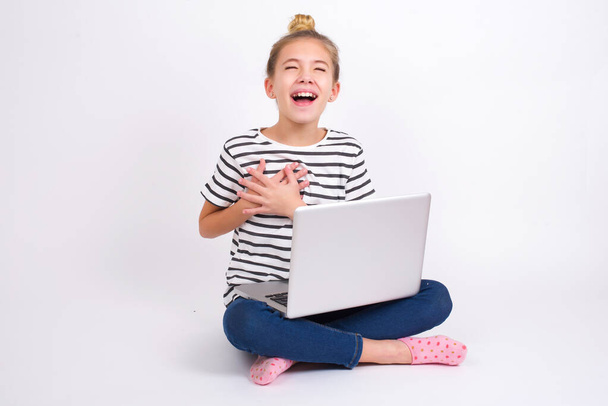 Beautiful caucasian teen girl sitting with laptop in lotus position on white background expresses happiness, laughs pleasantly, keeps hands on heart - Photo, Image