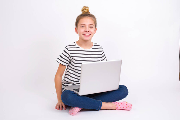 Beautiful caucasian teen girl sitting with laptop in lotus position on white background shows middle finger bad sign asks not to bother. Provocation and rude attitude. - Photo, Image