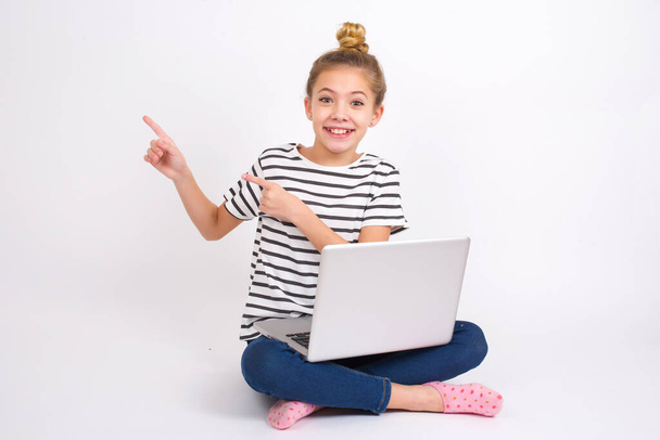 Beautiful caucasian teen girl sitting with laptop in lotus position on white background points at copy space indicates for advertising gives right direction - Foto, immagini