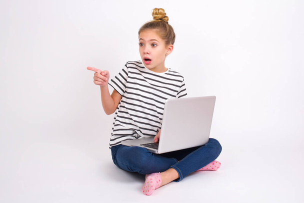 Oszołomiony piękny kaukaski nastolatek dziewczyna siedzi z laptopem w pozycji lotosu na białym tle z bardzo zaskoczony wyrażenie wskazuje daleko na przestrzeni kopii, wskazuje coś - Zdjęcie, obraz