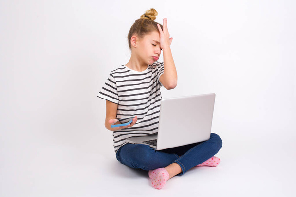 Upset depressed Beautiful caucasian teen girl sitting with laptop in lotus position on white background makes face palm as forgot about something important holds mobile phone expresses sorrow and regret blames - Foto, Imagem