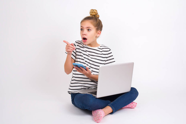Stunned Beautiful caucasian teen girl sitting with laptop in lotus position on white background points sideways right copy space, recommends product, sees astonishing thing - Photo, image