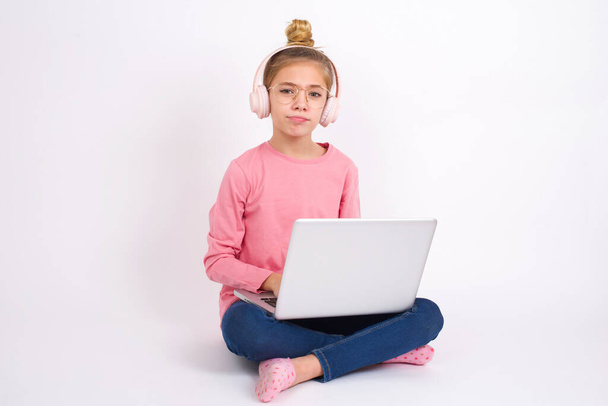 Mécontent Belle adolescente caucasienne assise avec un ordinateur portable en position lotus sur fond blanc fronce les sourcils visage se sent malheureux a quelques problèmes. Concept d'émotions et de sentiments négatifs - Photo, image