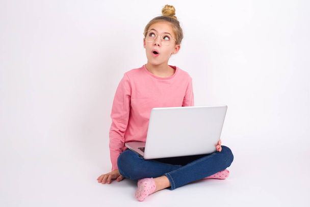 Sokkolt Gyönyörű kaukázusi tini lány ül laptoppal lótuszpozícióban fehér háttér néz üres tér nyitott szájjal sikoltozik: Ó, Istenem! Ezt nem hiszem el.. - Fotó, kép