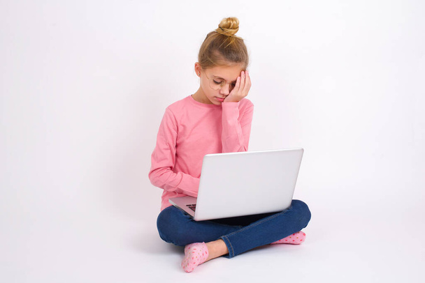 Schöne kaukasische Teenie-Mädchen sitzt mit Laptop in Lotus-Position auf weißem Hintergrund mit traurigem Gesichtsausdruck, der das Gesicht mit Händen bedeckt, während sie weint. Depressionskonzept. - Foto, Bild