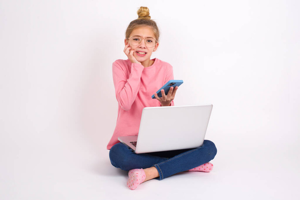 Bang grappig Mooie Kaukasische tiener meisje zitten met laptop in lotus positie op witte achtergrond holding telefoon en bijten nagels - Foto, afbeelding