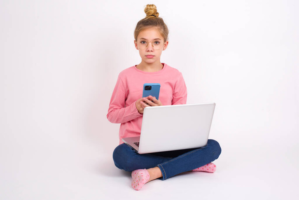 Portrait of serious confident Beautiful caucasian teen girl sitting with laptop in lotus position on white background holding phone in two hands - Fotografie, Obrázek