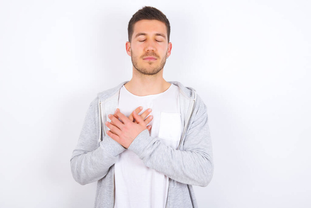 young caucasian man wearing casual clothes over white background closes eyes and keeps hands on chest near heart, expresses sincere emotions, being kind hearted and honest. Body language and real feelings concept. - Photo, Image