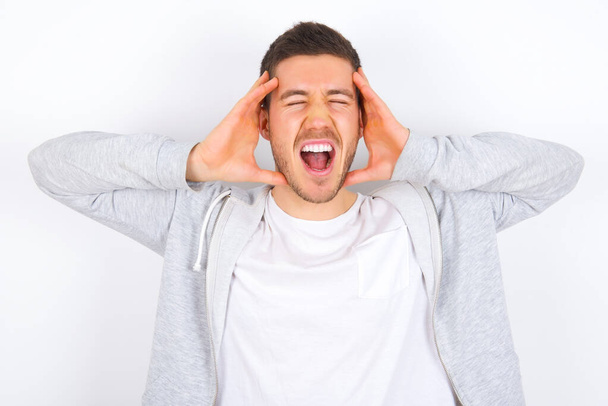 Shocked panic young caucasian man wearing casual clothes over white background holding hands on head and screaming in despair and frustration. - Foto, immagini