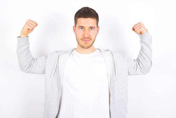 Waist up shot of young caucasian man wearing casual clothes over white background raises arms to show muscles feels confident in victory, looks strong and independent, smiles positively at camera. Sport concept. - Foto, Imagen