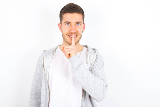Smiling young caucasian man wearing casual clothes over white background makes shush gesture, holds fore finger over lips hides secret. Be mute, please. - Foto, immagini