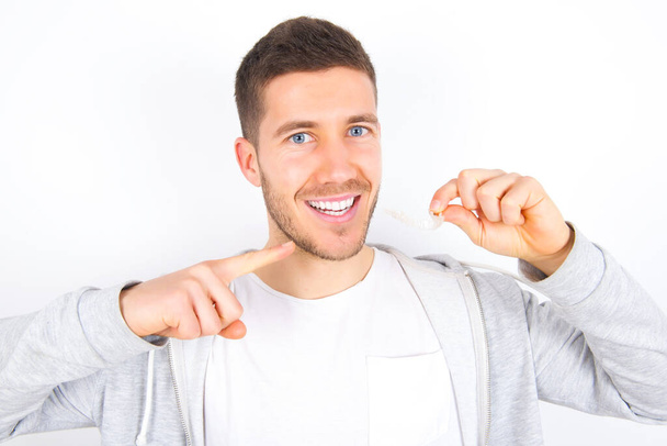 young caucasian man wearing casual clothes over white background holding an invisible aligner and pointing to her perfect straight teeth. Dental healthcare and confidence concept. - Foto, Imagem