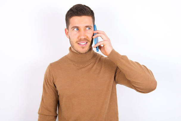 Pleasant looking happy young caucasian man wearing turtleneck over white background has nice telephone conversation and looks aside, has nice mood and smiles positively while talks via cell phone - Photo, image