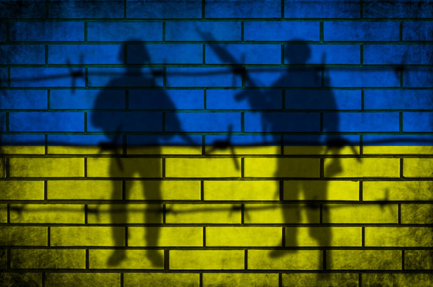 Ukraine flag and military background. Barbed fence and armed men silhouette. Conflict and war concept. Grunge texture on brick wall - Photo, Image