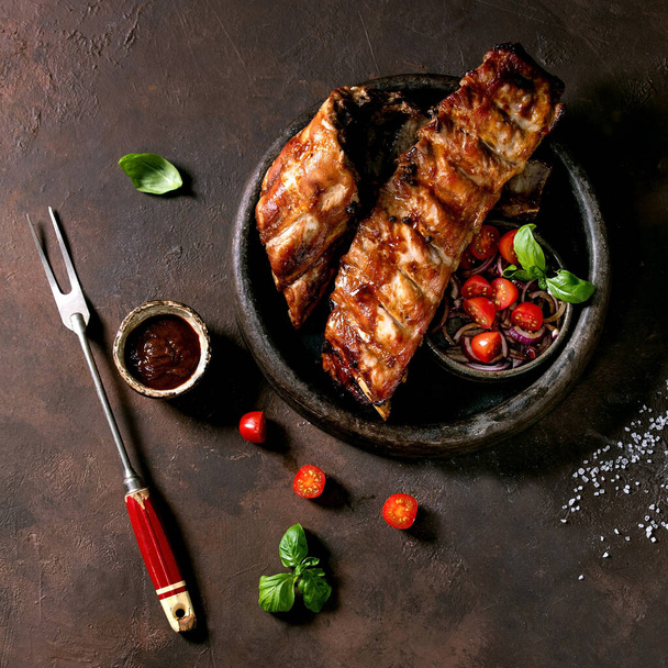 Grilled pork bbq ribs in ceramic plate served with meat fork, vegetable salad, cherry tomatoes, basil and barbeque sauce over dark brown texture background. Top view, flat lay. Square image - Φωτογραφία, εικόνα