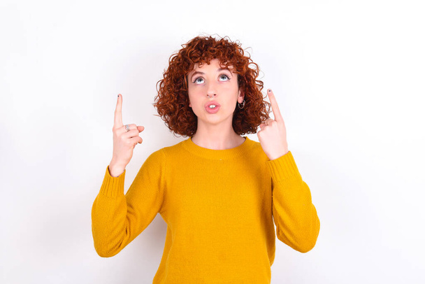 young redhead girl wearing yellow sweater over white background   being amazed and surprised looking and pointing up with fingers showing something strange. - Foto, afbeelding