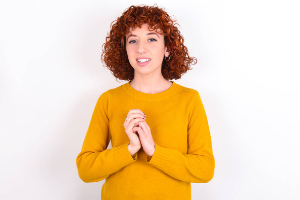 Dromerig charmant jong roodharig meisje dragen gele trui over witte achtergrond met aangename uitdrukking, houdt de handen bij elkaar enthousiast over iets aangenaams. - Foto, afbeelding