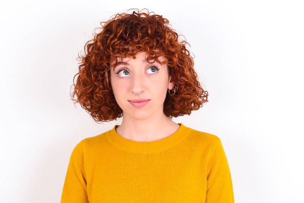 young redhead girl wearing yellow sweater over white background   has worried face looking up,  lips together, being upset thinking about something important, keeps hands down. - Photo, Image