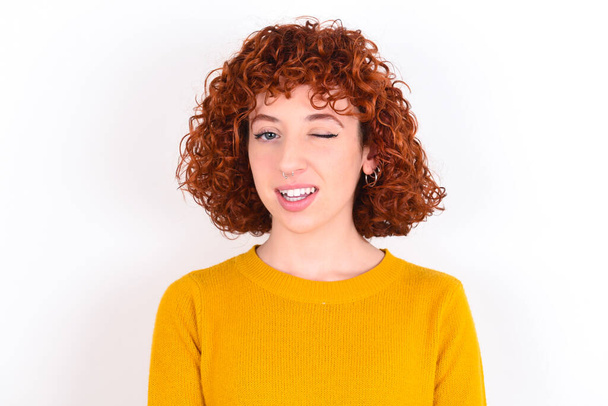 young redhead girl wearing yellow sweater over white background   blinking eye with pleasure having happy expression. Facial expressions and people emotions concept. - Fotó, kép