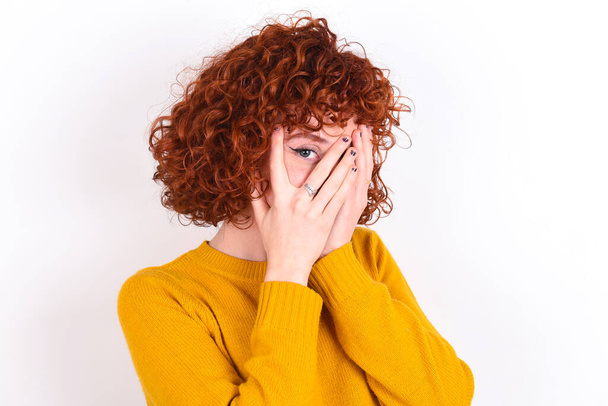 young redhead girl wearing yellow sweater over white background   covering face with hands and peering out with one eye between fingers. Scared from something or someone. - Foto, immagini
