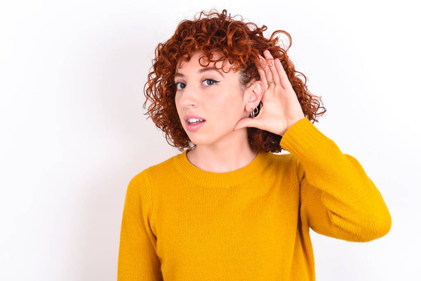Oh my God! Funny astonished young redhead girl wearing yellow sweater over white background   opening mouth widely looking aside, with hand near ear trying to listen to gossips. - Photo, Image