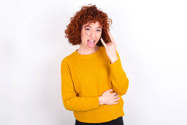 Retrato de uma jovem ruiva louca vestindo suéter amarelo sobre fundo branco mostrando chifres de língua até gesto, expressando emoção de estar em concerto da banda. - Foto, Imagem