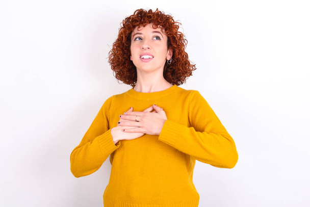 Happy smiling young redhead girl wearing yellow sweater over white background   has hands on chest near heart. Human emotions, real feelings and facial expression concept. - Photo, Image